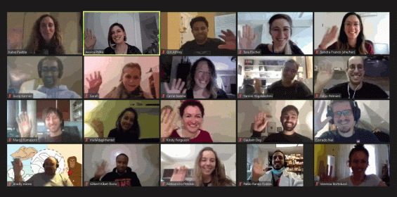 A grid of sixteen video call participants smiling and waving at their cameras. Each participant is in a separate window, with different backgrounds and lighting, creating a friendly and diverse atmosphere.