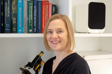 A person with short blonde hair and a black shirt smiles at the camera. They stand in front of shelves with books and a microscope.