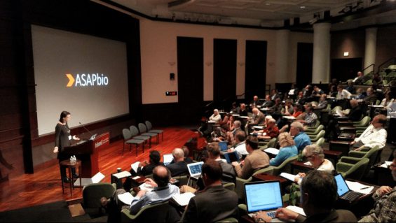 A speaker presents at an ASAPbio conference in an auditorium. Attendees are seated, using laptops and taking notes. The room is well-lit, with a large screen displaying the ASAPbio logo.