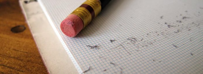 A black pencil with a red eraser rests on a spiral grid notebook. Eraser shavings are scattered on the open page, and the wooden surface underneath is visible.