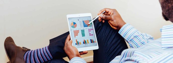A person in business attire sits cross-legged, holding a tablet. The screen displays colorful charts and graphs. The person is using a stylus to interact with the tablet. The setting appears to be a casual office.