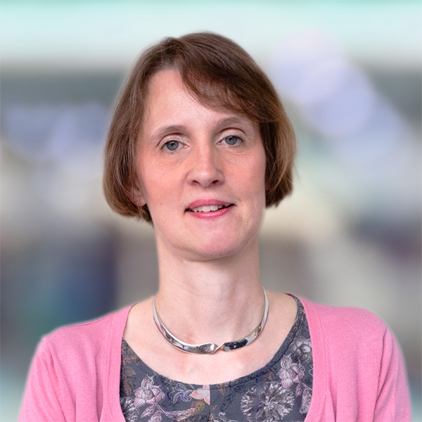 A woman with short brown hair, wearing a pink cardigan, patterned top, and silver necklace. She is smiling slightly with a blurry, light background behind her.