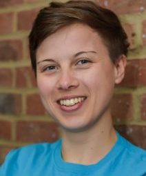 A person with short hair smiles at the camera, wearing a blue shirt. They are standing against a brick wall.