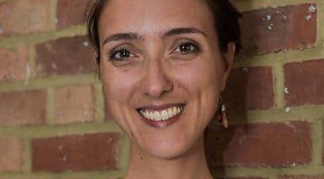 Smiling person with short hair, wearing a patterned shirt and earrings, stands against a brick wall.