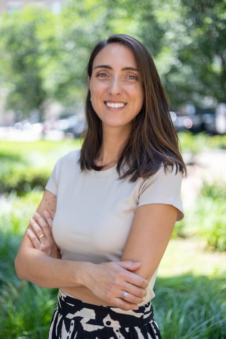 A woman with long brown hair, wearing a light beige shirt and patterned black skirt, stands outdoors. She smiles with her arms crossed, with lush green trees and grass in the background.
