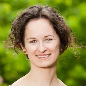 A person with curly dark hair smiles at the camera with a blurred green background of foliage.