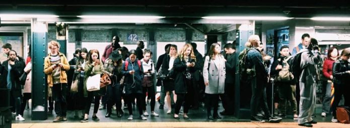A crowded subway platform with a diverse group of people waiting. Many are holding or looking at their phones. The platform is dimly lit, and a track is visible in the foreground. The atmosphere is busy and typical of a city commute.