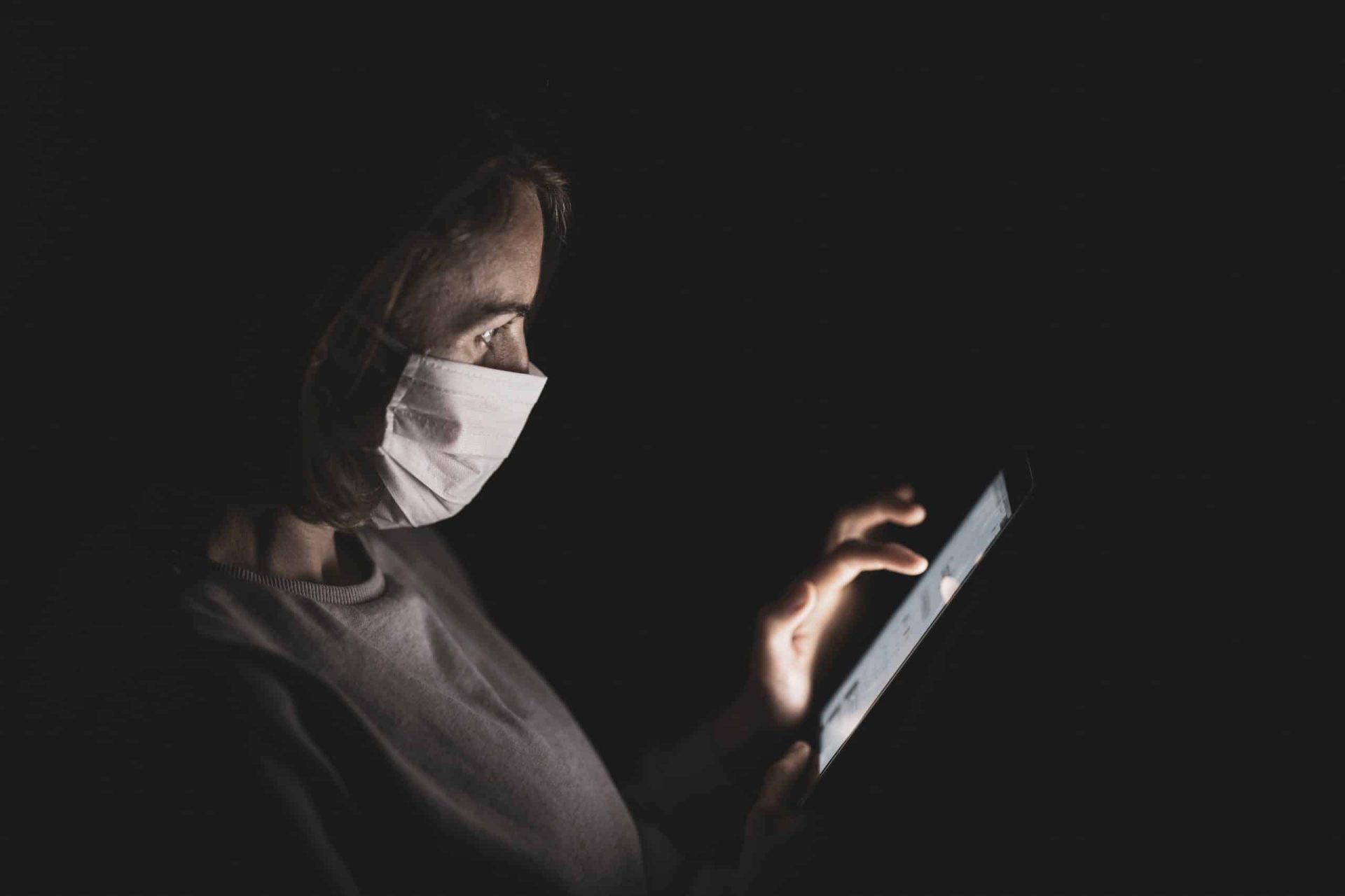 A person wearing a mask is illuminated by the glow of a tablet in the dark. They are looking intently at the screen while touching it with their fingers.