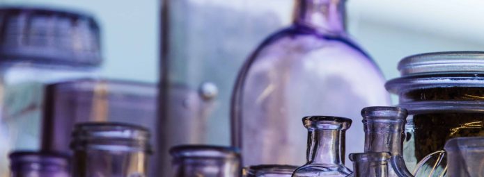 A collection of vintage glass bottles, in varying shades of purple, are clustered together. The bottles differ in shape and size, with some having narrow necks and others with wider openings, set against a softly lit background.