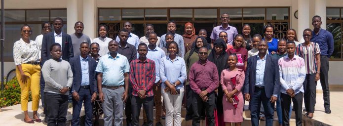 A diverse group of people stands outside a building, posing for a group photo on steps. They are dressed in a mix of formal and casual attire, smiling at the camera. The background features large windows and a door.