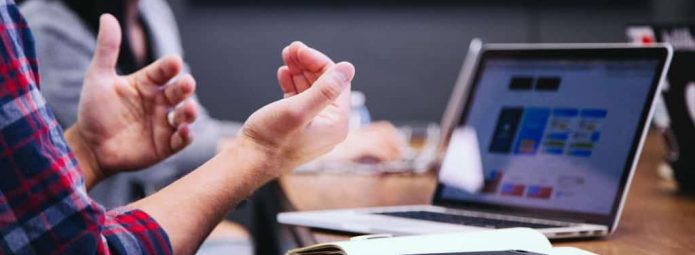 A person wearing a plaid shirt gestures with their hands while sitting at a table with a laptop and notebook. Another person is blurred in the background. The laptop screen displays colorful graphics.