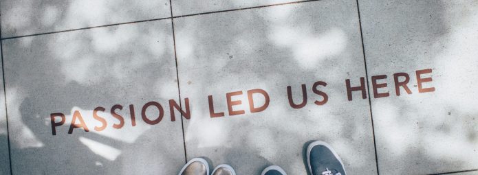 Looking down at the feet of two people standing on a sidewalk. The sidewalk is stamped with the words "passion led us here."