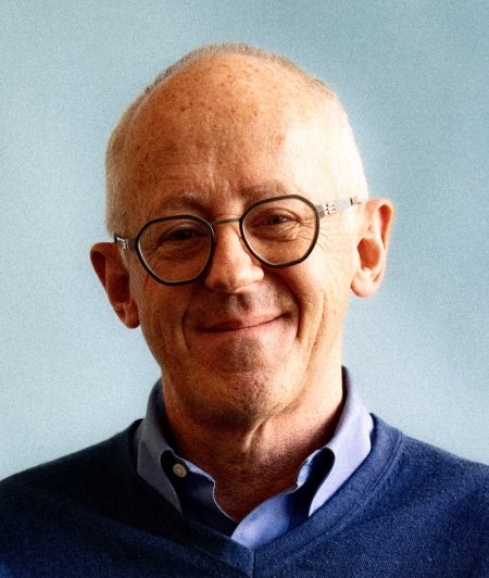 Smiling older person with glasses, short light hair, wearing a blue sweater over a collared shirt, against a pale blue background.