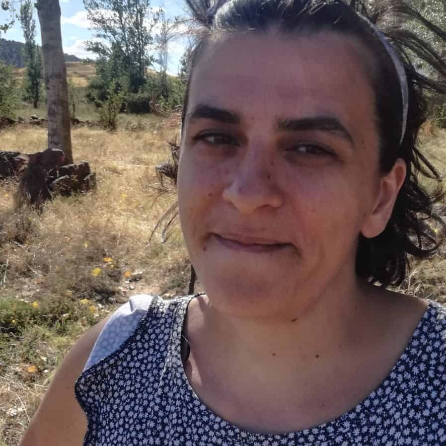 Woman with dark hair, wearing a black and white patterned sleeveless top, stands outdoors in a sunny, rural landscape. There are trees and dry grass in the background.