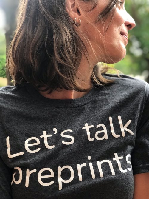 A woman with shoulder-length brown hair is smiling and looking to the side. She is wearing a dark gray t-shirt with the text Lets talk preprints printed in white letters. The background is blurred greenery.