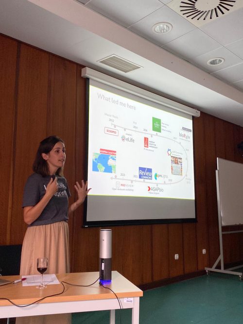 A person is giving a presentation in a room with wood-paneled walls. They are standing in front of a projector screen displaying a timeline with various logos and text. A table with a wine glass and a small device is in the foreground.