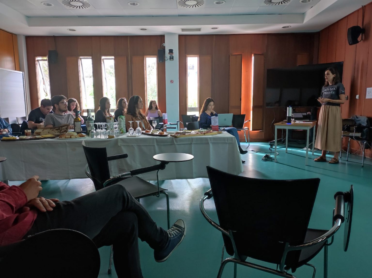 A person stands presenting to a seated audience in a room. The audience observes a table with snacks and drinks in the background. A large screen and whiteboard are visible, and natural light filters through tall windows.