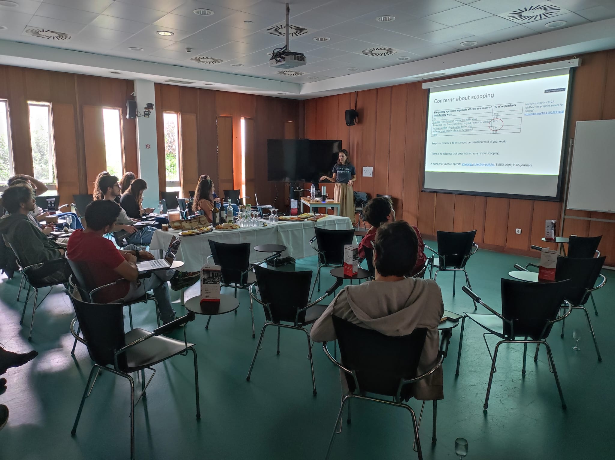 A person stands in front of a projected presentation in a conference room, speaking to an audience sitting in a semi-circle. The room has wooden walls and a large television. Tables with refreshments are at the center.