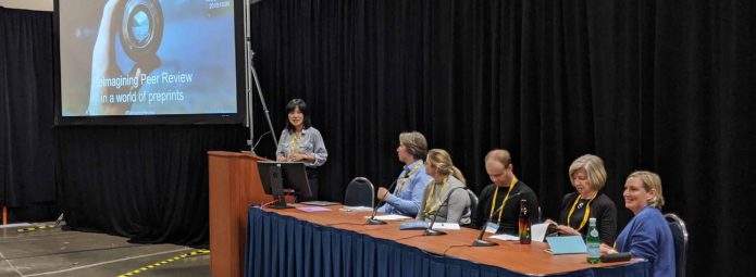 A woman stands at a podium presenting a slideshow titled Reimagining Peer Review in a World of Preprints. Five people sit at a table on stage, listening. The setting is a conference with black curtains and exhibition hall lighting.