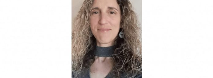 Person with long curly hair wearing a gray top, smiling slightly, and standing against a plain background.