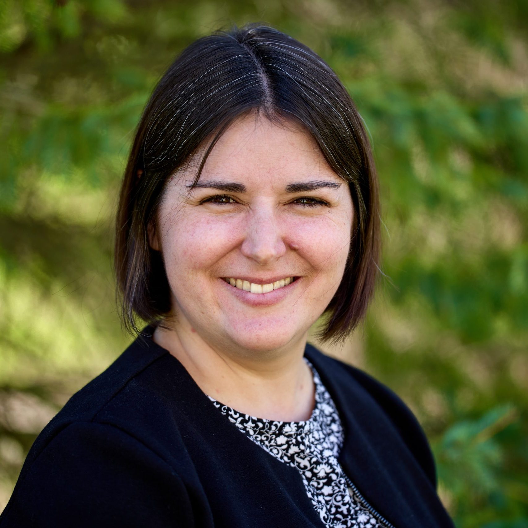 A person with short dark hair is smiling, wearing a black jacket and a patterned top. The background is lush and green, suggesting an outdoor setting with trees.