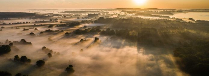 Aerial view of a misty landscape at sunrise. The sun casts long shadows over fields and clusters of trees, with a winding road visible. The sky is a soft gradient of orange and blue, enhancing the serene atmosphere.