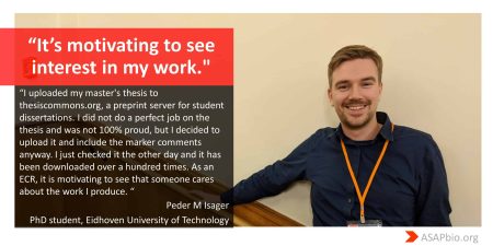 A person with short hair and a beard is smiling while wearing a dark blue shirt with an orange lanyard, standing against a plain light-colored wall.
