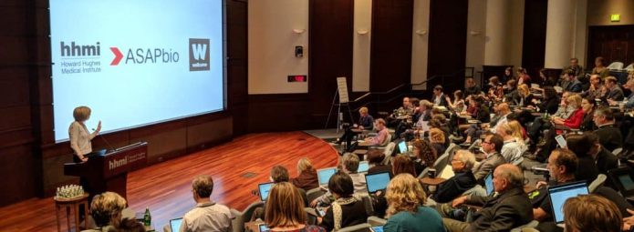 A speaker presents at a conference in a lecture hall filled with attendees. The large screen displays logos of Howard Hughes Medical Institute, ASAPbio, and the Wellcome Trust. Audience members are seated with laptops open, attentively listening.