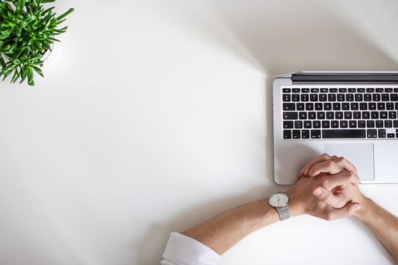 A person with folded hands rests on a white desk beside a silver laptop. A small green plant is situated in the top left corner of the desk, creating a minimalist and organized workspace setting.