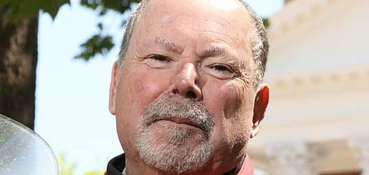 A man with a short gray beard and mustache is wearing a red and black jacket. He is outdoors, standing near trees, with a building visible in the background.
