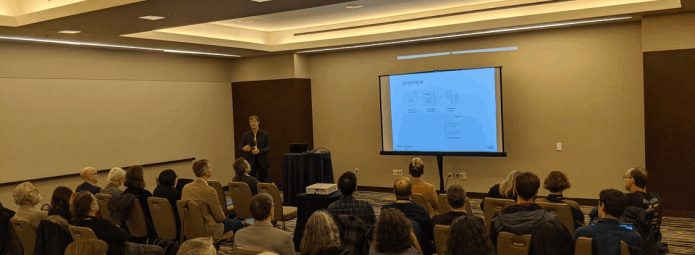 A person is presenting a slideshow to an audience in a conference room. The audience sits in rows of chairs, facing the presenter and a large screen displaying a diagram. The room has bright overhead lighting.