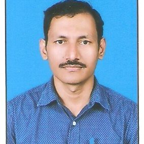 Portrait of a person with short black hair and a mustache, wearing a blue patterned shirt. The background is a solid blue color.