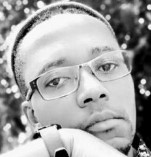 A black and white close-up photo of a person wearing glasses, resting their chin on their hand. The background is softly blurred with a hint of foliage, conveying a contemplative mood.