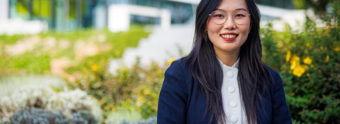 Photograph of Victoria Yan wearing a white sweater and navy blazer in front of a landscaped campus, with a glass building in the background
