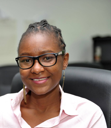 A person with short braided hair, wearing glasses and a light pink shirt, smiles while sitting in an office chair. The background is blurred, featuring a desk and office equipment.