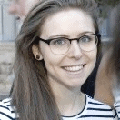 A person with long hair and glasses smiles at the camera. They are wearing a striped shirt. There are blurred figures and a building in the background.