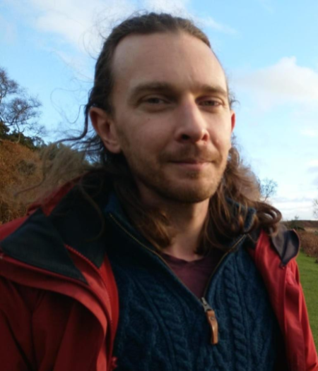 A person with long hair stands outdoors, wearing a red jacket and blue sweater. The background shows clear skies and greenery, suggesting a natural setting.