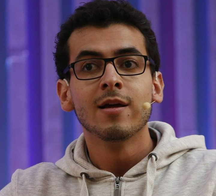 A person with short dark hair and glasses is speaking into a microphone. They are wearing a light gray hoodie and standing in front of a blue and purple striped background.