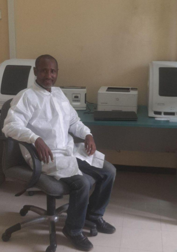 A man in a white lab coat sits on an office chair in a room with beige walls. Behind him, there are two computer monitors and a printer on a desk. He looks at the camera with a neutral expression.
