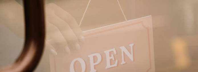 A hand is seen through a glass door, turning an OPEN sign to face outward, indicating that the establishment is open. The sign is rectangular with a simple border.