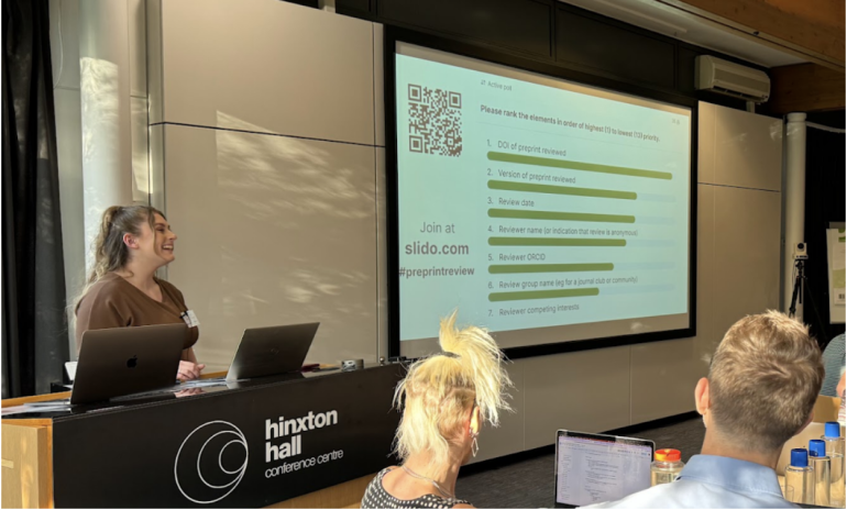 A presenter stands at a podium next to a projection screen in a conference room. The screen displays a list with a QR code and text discussing review criteria. Attendees are seated, some with laptops open. The room has a sign reading hinxton hall conference centre.